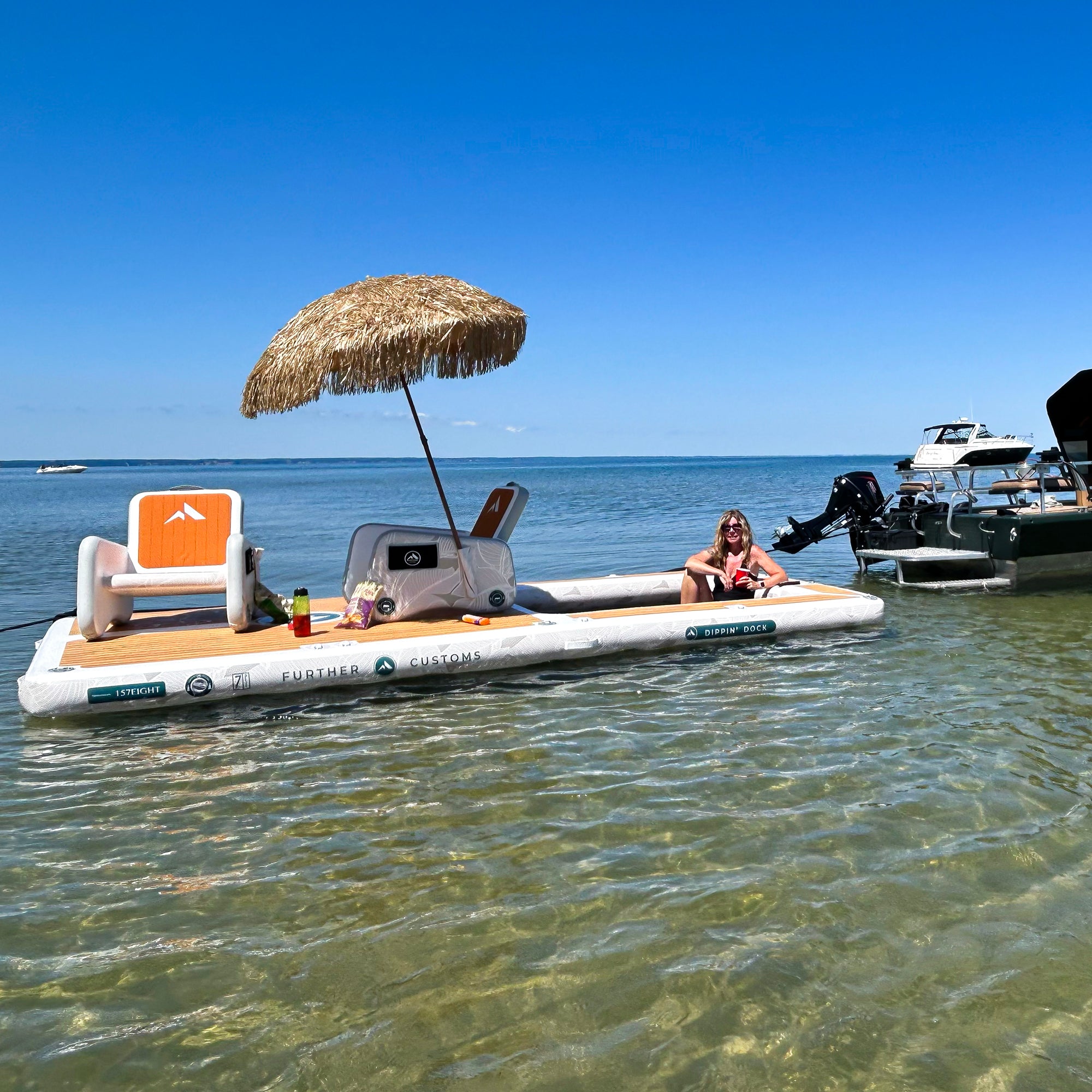 A white and tan inflatable floating lounge platform on shallow blue water, with a straw-style thatched umbrella, two captains inflatable boat chairs bote, and a woman sitting on the mesh water hammock. A boat is moored nearby under a clear blue sky and anchored adjacent.